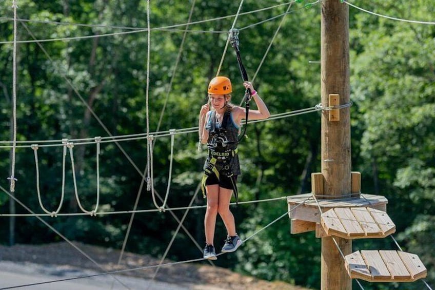 A person on a rope course with wooden platforms and a forested background. 1