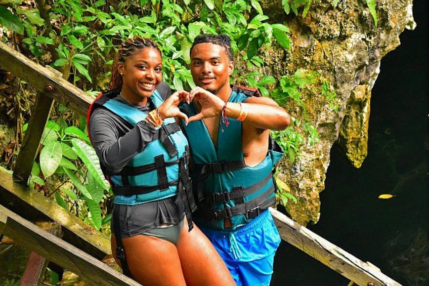 Two people making a heart shape with their hands, wearing life jackets, standing on a wooden bridge with greenery in the background. 1