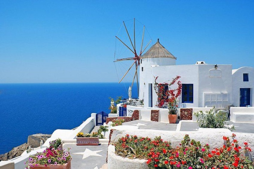 A white building with a windmill, red flowers, and the sea in the background. 3