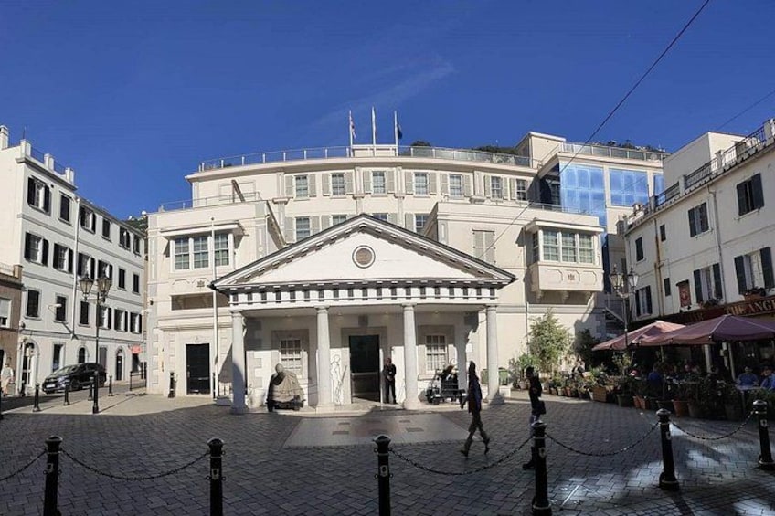 Un bâtiment historique à la façade classique, une place pavée et une clôture en treillis métallique. 2