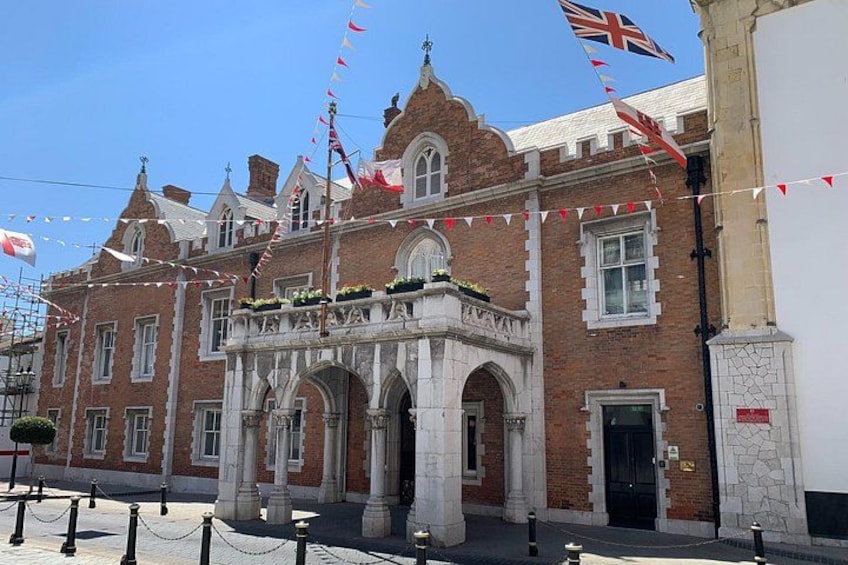 A historic building with a prominent entrance, red brick walls, and white stone detailing. Flags are flying above the entrance, and there are decorative elements like flower boxes and a small balcony. 1