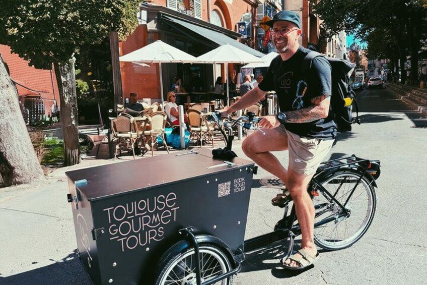A man on a bicycle with a box labeled 'TOUJOUSE GOURMET TOURS' on the sidewalk. 4