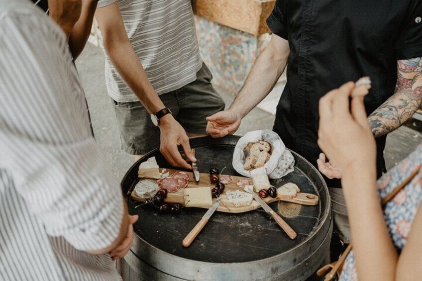 A group of people around a barrel with various cheeses and meats. 5
