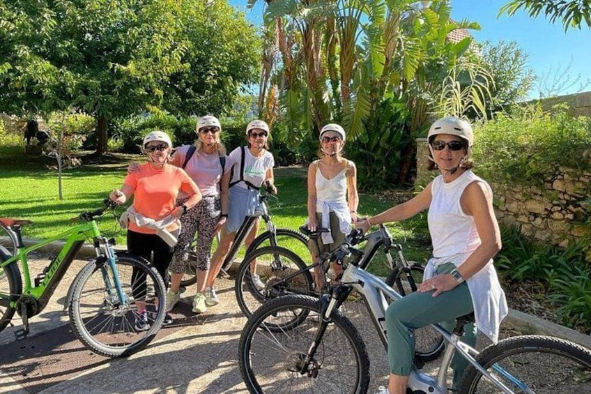 A group of people on bicycles in a park with greenery and a stone wall in the background. 5