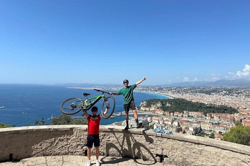 Two people performing a bicycle stunt on a cliff overlooking a coastal city. 4