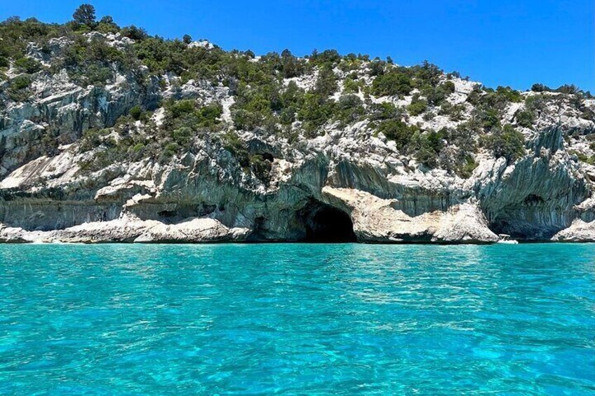 A coastal landscape with clear turquoise waters, rocky cliffs, and a cave entrance. 4