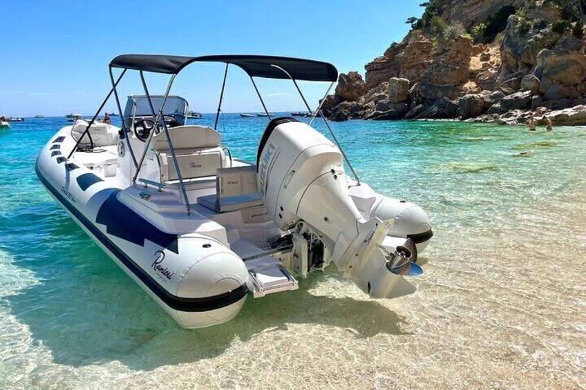 A white motorboat with a canopy, docked at a clear, shallow coastal area with rocky cliffs in the background. 5