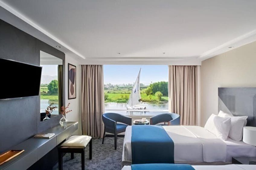 A hotel room with a bed, a desk, a chair, and a view of a lake with a sailboat. 5