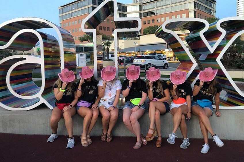 A group of people sitting on a bench in front of a large, colorful sign with the word 'ALEX' on it. 5