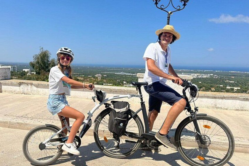 Twee personen op fietsen, één met een witte helm en de ander met een strohoed, op een verhard pad met een schilderachtig uitzicht op een kustlandschap. 5