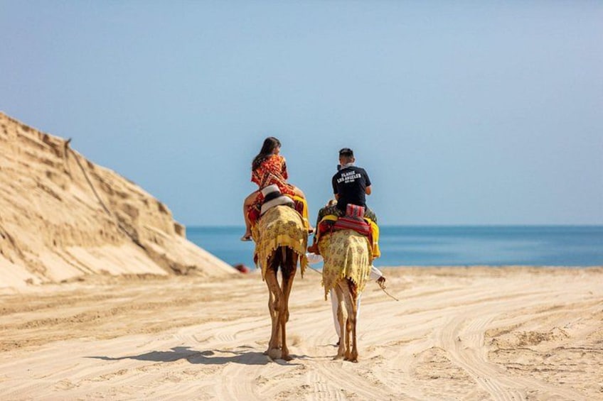 Two people riding camels on a sandy path near a body of water. 1