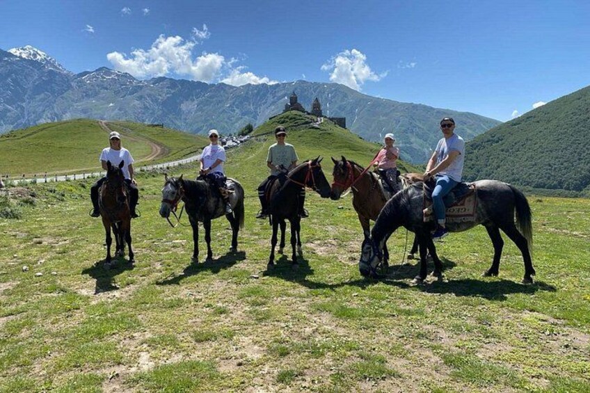 Four people riding horses on a grassy hillside with mountains in the background. 1