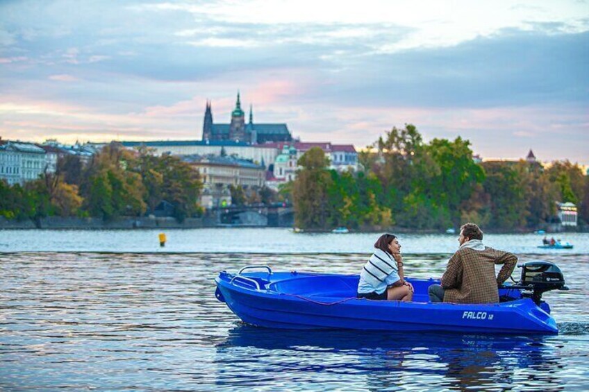 Two people in a blue boat on a river with a cityscape in the background. 1