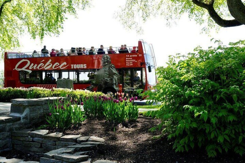 A red double-decker bus with 'Quebec Tours' written on it, surrounded by greenery and a statue. 3