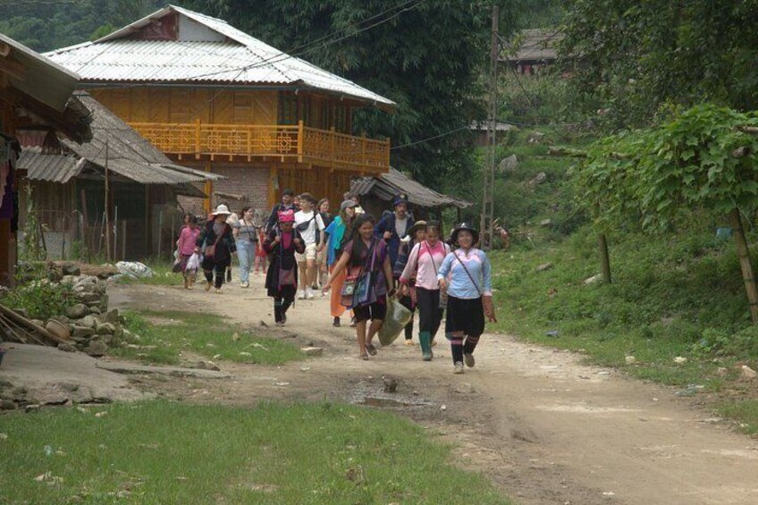 A group of people walking on a dirt path in a rural area with buildings and greenery. 1