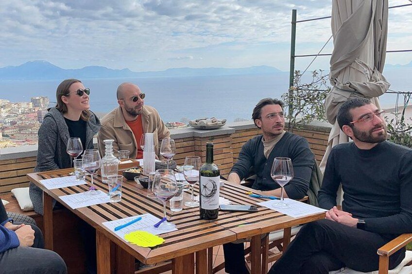 A group of people seated at a wooden table outdoors with a scenic view of the sea and mountains in the background. 4