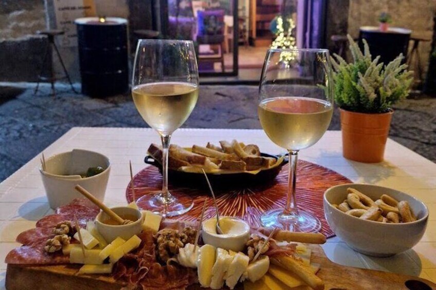 En bricka med olika sorters charkuterier, ostar och kex, tillsammans med två glas vitt vin, placerad på ett bord utomhus. 1