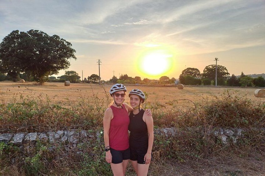 Two people wearing helmets and cycling attire standing in a field with hay bales and a stone wall in the foreground, and a sunset in the background. 3
