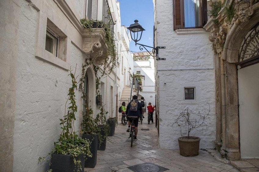 A narrow street with white buildings, potted plants, and a cyclist in the distance. 2