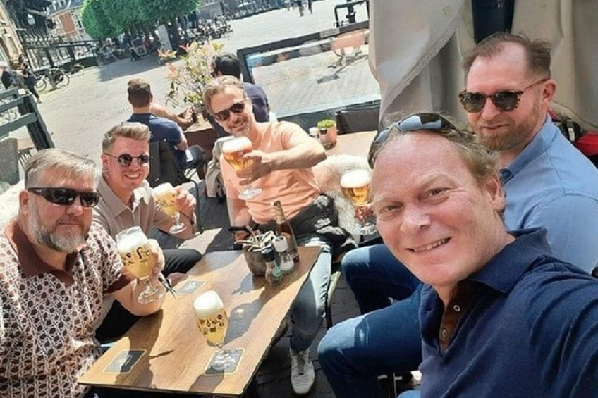 A group of people sitting at an outdoor table, holding drinks, with a city street in the background. 2