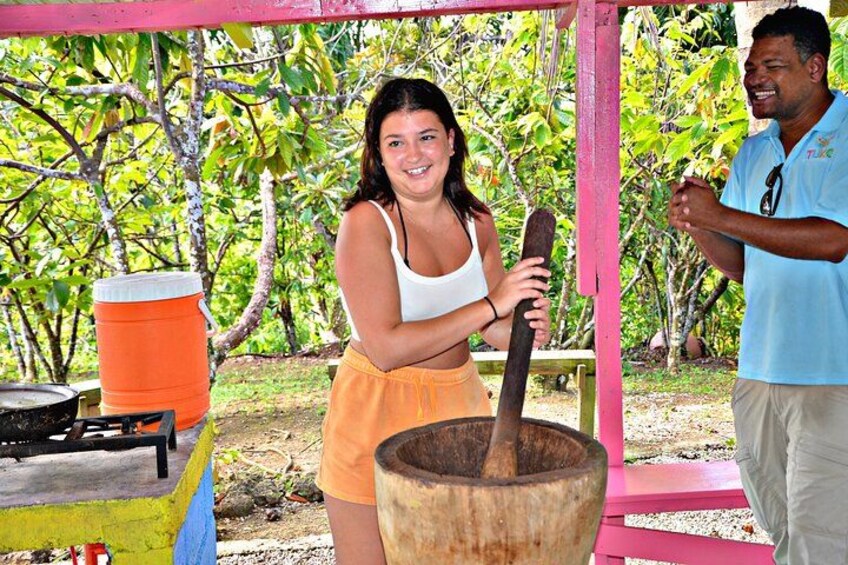 A person using a large wooden tool, possibly for cooking or preparing food, in an outdoor setting with trees and a pink structure. 3