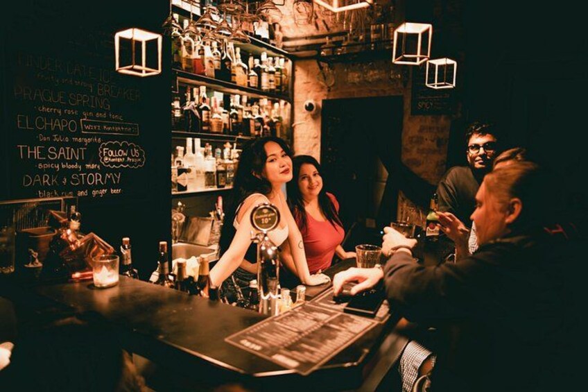 Un bar con un menú en pizarra que enumera bebidas, un bartender atendiendo a un cliente, y un interior tenuemente iluminado con luces colgantes. 3