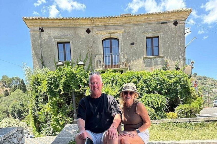 A man and a woman sitting on a bench in front of a stone building with a balcony, surrounded by greenery. 2
