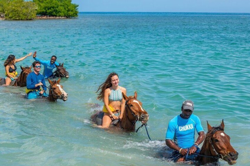 People riding horses in shallow water near a beach with clear skies. 5