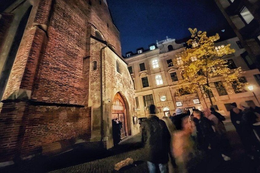 Ein historisches Gebäude mit Ziegel-Fassade und Bogenportal, umgeben von modernen Gebäuden und einem Baum. 2