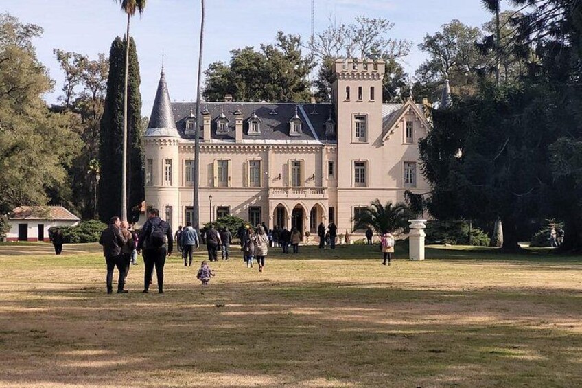 A large, historic building with a tower and arched windows, surrounded by a grassy area with people walking. 1