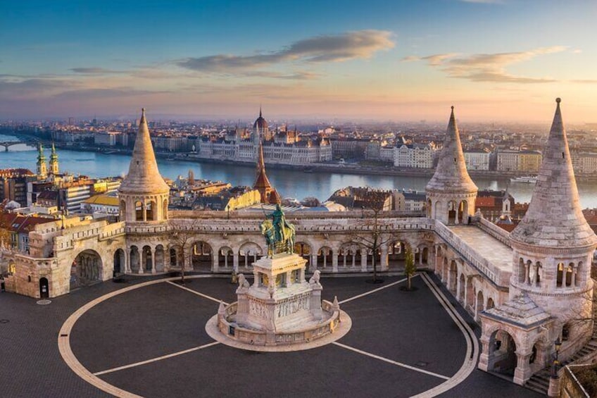 Una plaza histórica con un monumento central, rodeada de edificios con tejados puntiagudos y un río al fondo. 1