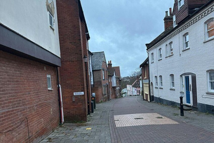 A narrow street lined with red brick and white buildings, a paved area with a patterned design, and a blue door on the right. 2