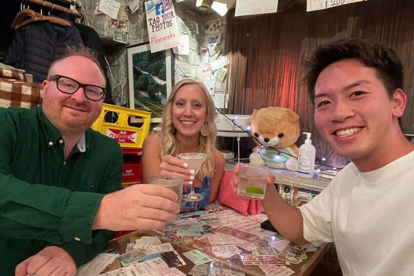Three people sitting at a table with drinks, surrounded by various items and a teddy bear. 2
