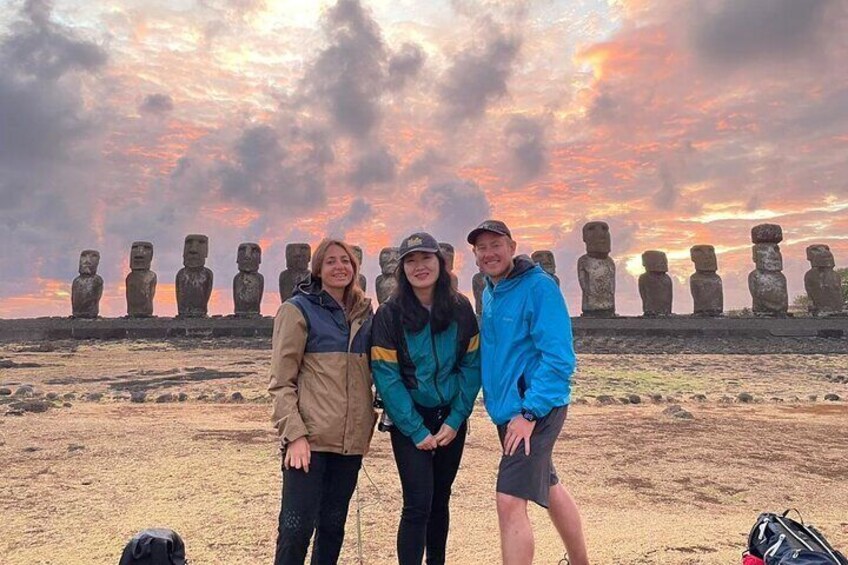Three people standing in front of a row of large stone statues under a cloudy sky. 2