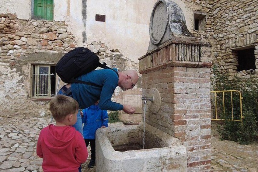 A man with a backpack leaning over a stone well, a child in a red hoodie watching, and a stone building with a green window in the background. 1