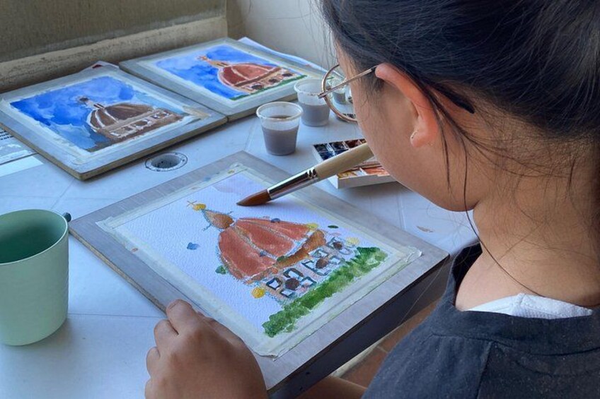A person painting on a canvas with a brush, a cup, and paint tubes on a table. 2