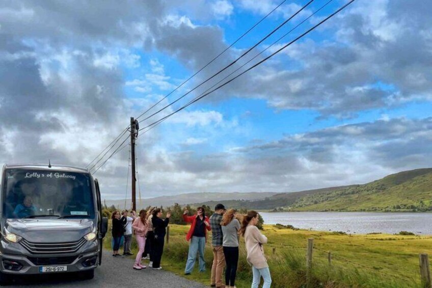 Een groep mensen staat langs een weg, met een bus met de tekst 'Lolly's of Galley' en een meer op de achtergrond. 2