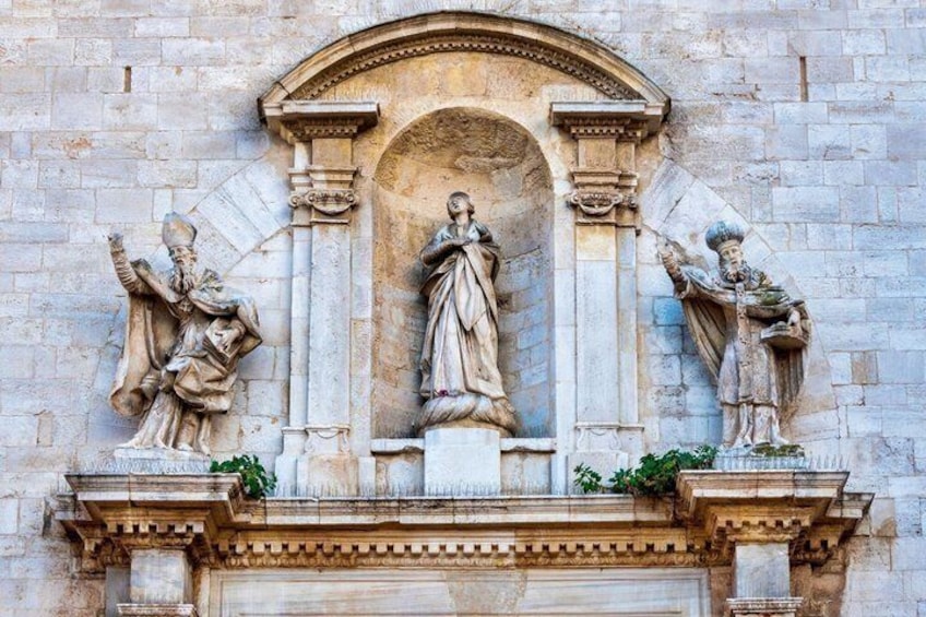 A stone facade with a central archway, flanked by statues of figures holding objects. 2