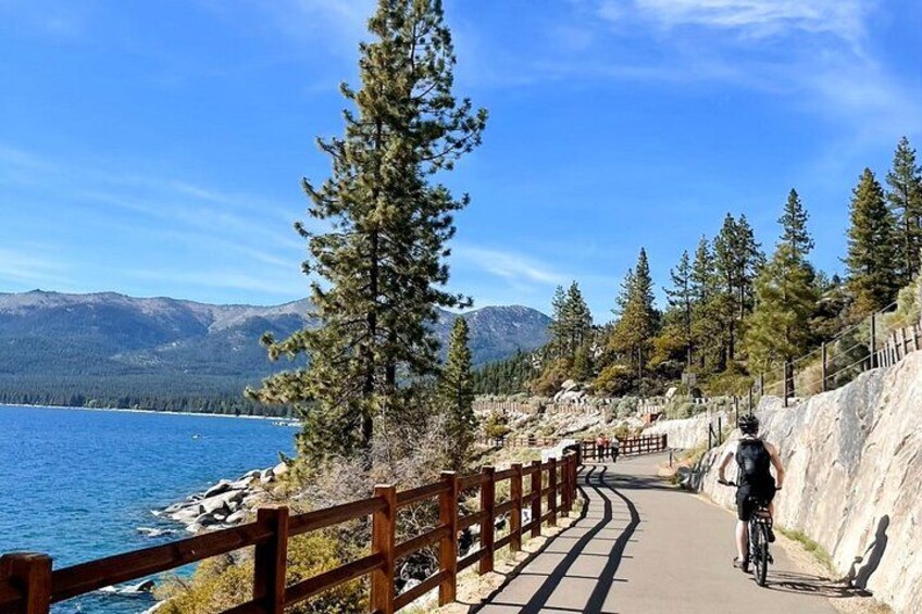 Un camino pavimentado junto a un lago con una cerca de madera, rodeado de árboles y montañas al fondo. 1