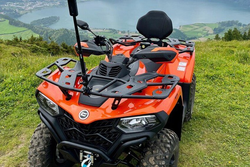 An orange ATV with a black seat and storage compartment, parked on grass with a scenic view of a lake and hills in the background. 3