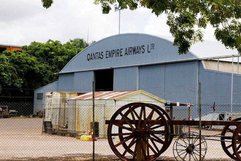 Un grande edificio industriale con un cartello che recita 'Qantas Empire Airways Ltd.' davanti a un'area recintata con una grande ruota di legno. 3