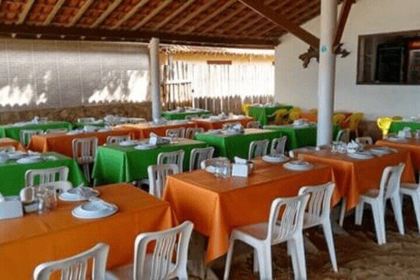An outdoor dining area with tables covered in orange and green tablecloths, white plastic chairs, and a rustic wooden ceiling. 5