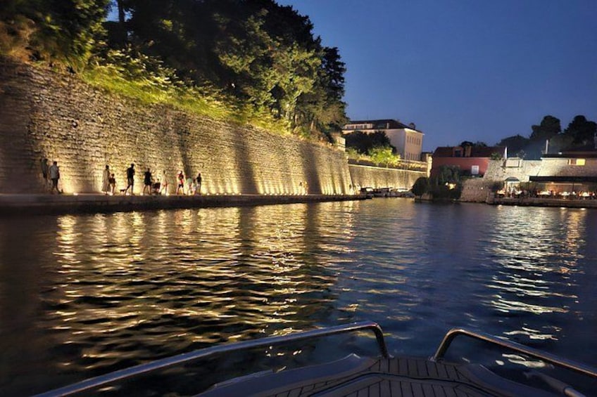 A river with people walking along the embankment, a boat docked at the pier, and buildings in the background. 2