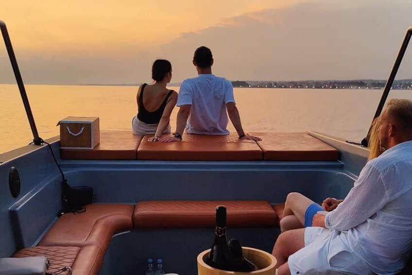 Un barco con dos personas sentadas en la cubierta, una tercera persona en la cubierta inferior, y un atardecer al fondo. 3