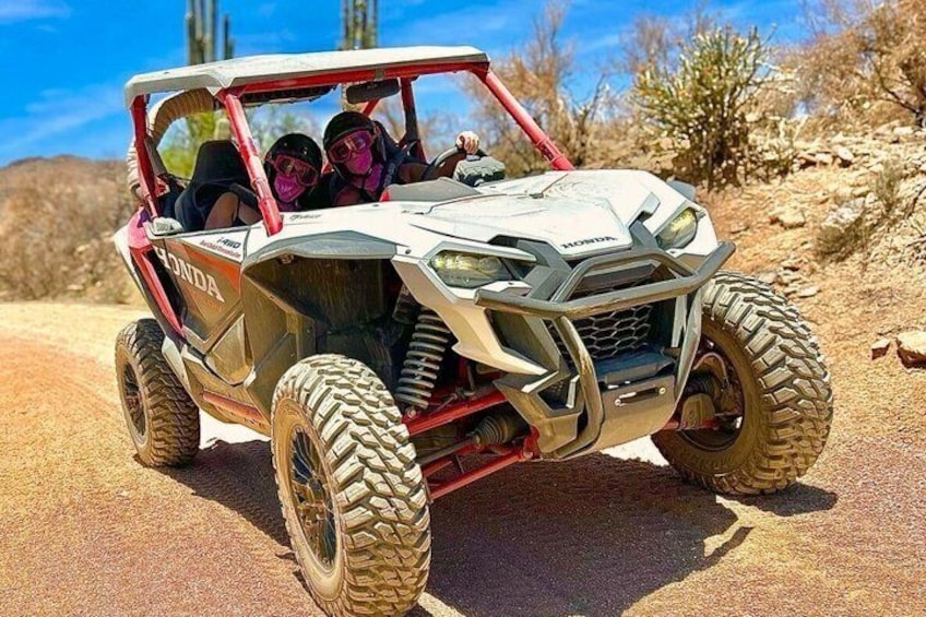 A white off-road vehicle with red accents, a driver, and a passenger, on a dirt path with sparse vegetation and rocks. 1