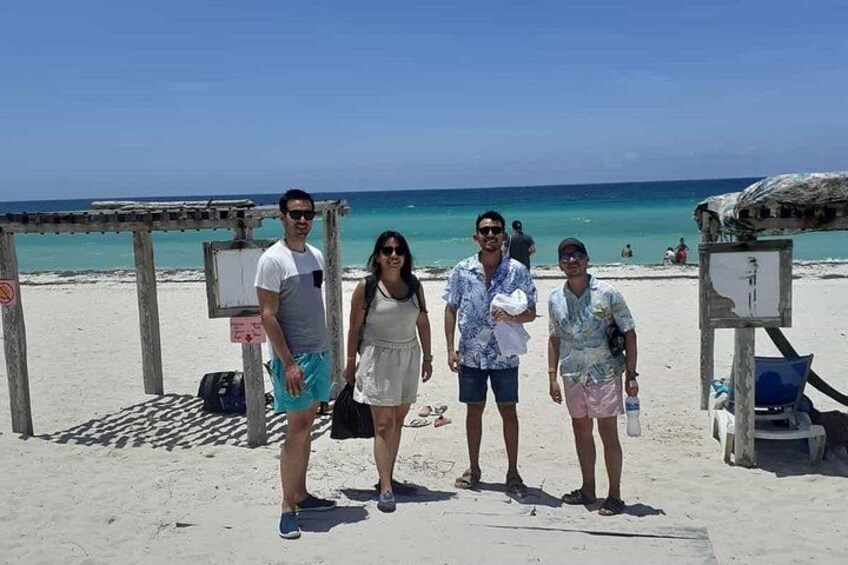 A beach with people standing under a wooden structure, a clear blue sky, and the ocean in the background. 2