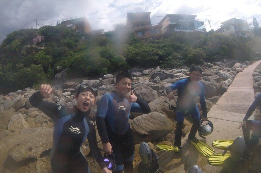 Three people in wetsuits near rocky terrain with a pathway and buildings in the background. 1