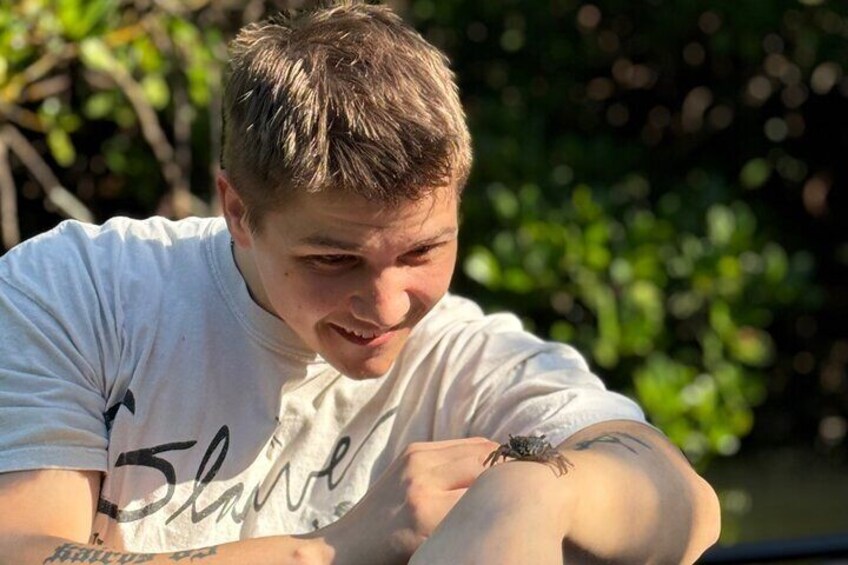 A person wearing a white t-shirt with a tattoo on their arm, sitting outdoors with greenery in the background. 3