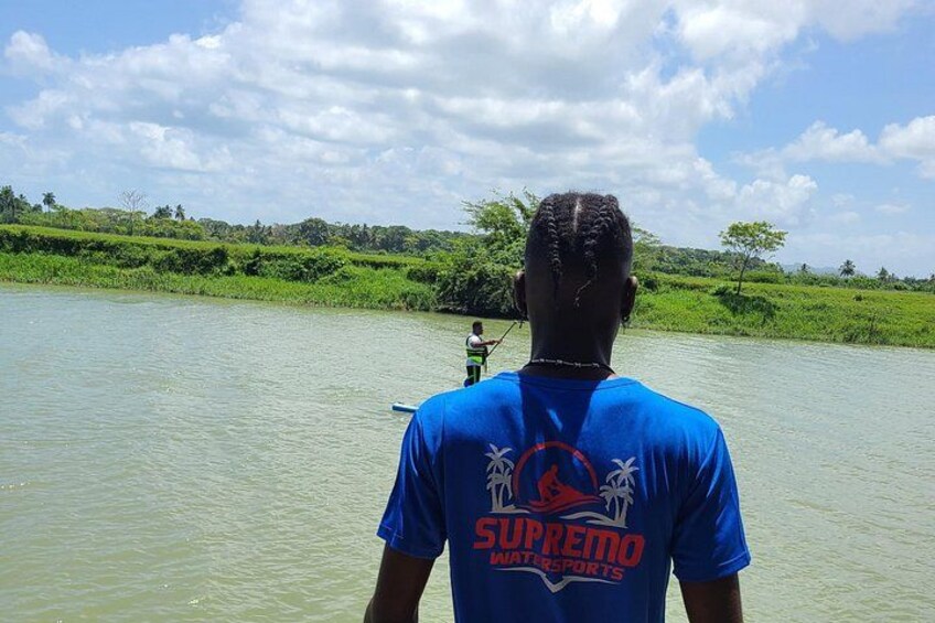 A person in a blue shirt with 'SUPREMO WATERSPORTS' on the back, standing in a body of water with a green landscape in the background. 5