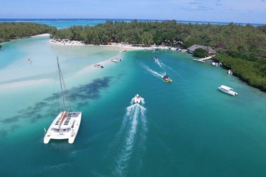 Un mar turquesa y transparente con barcos, un velero y un yate. Se observa una playa con edificios y una zona boscosa al fondo. 1